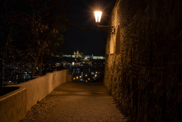 вид через старую подсвеченную каменную стену XVII века и свет уличного освещения, а на заднем плане - подсвеченный замок и церковь Святого Вита, река Влтава в ночное время - Фото, изображение
