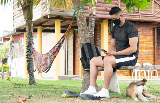 Latin man using the cell phone, wearing a black mask, outdoor with his pet. - Photo, Image