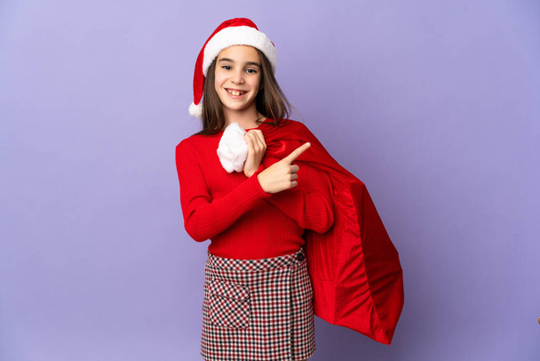 Menina com chapéu e saco de Natal isolado no fundo roxo apontando para o lado para apresentar um produto - Foto, Imagem
