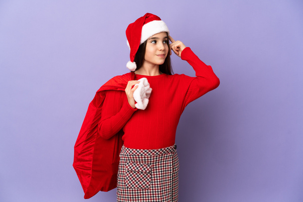 Kleines Mädchen mit Hut und Weihnachtssack isoliert auf lila Hintergrund mit Zweifeln und Denken - Foto, Bild
