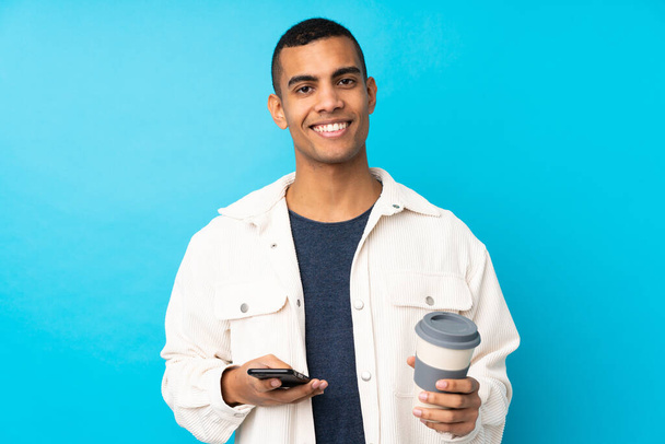 Jovem afro-americano sobre fundo azul isolado segurando café para levar e um celular - Foto, Imagem