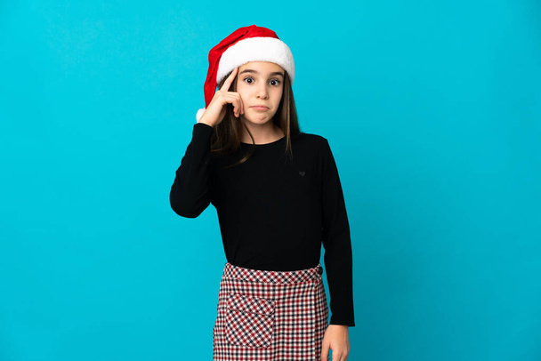 Маленька дівчинка з різдвяним капелюхом ізольована на синьому фоні думаючи про ідею
 - Фото, зображення