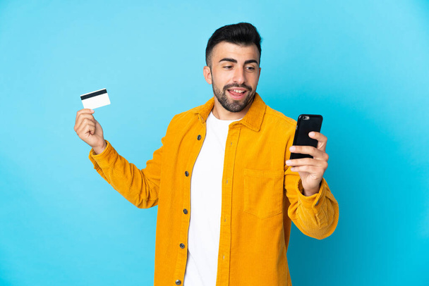 Caucasian man over isolated blue background buying with the mobile with a credit card - Fotoğraf, Görsel
