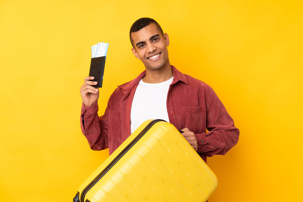 Молодой афроамериканец с изолированным желтым фоном в отпуске с чемоданом и паспортом - Фото, изображение