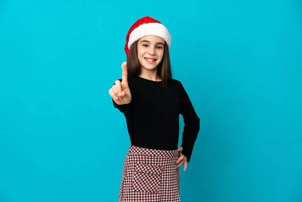 Petite fille avec chapeau de Noël isolé sur fond bleu montrant et levant un doigt - Photo, image