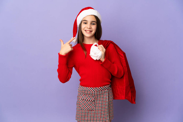 Little girl with hat and Christmas sack isolated on purple background giving a thumbs up gesture - Photo, Image