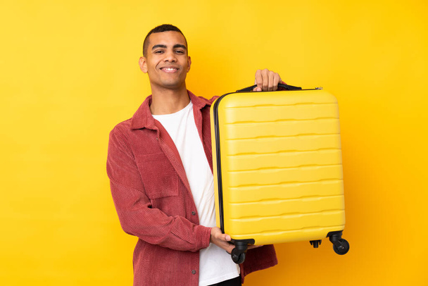 Νεαρός Αφροαμερικανός σε απομονωμένο κίτρινο φόντο σε διακοπές με ταξιδιωτική βαλίτσα - Φωτογραφία, εικόνα