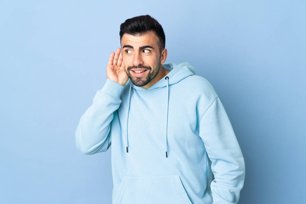 Caucasian man over isolated blue background listening to something by putting hand on the ear - Foto, Imagem