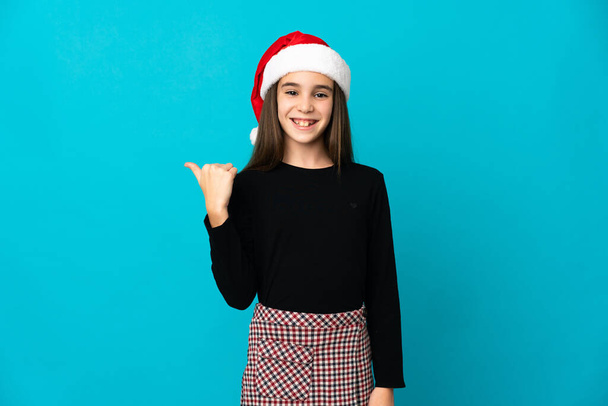 Mavi arka planda izole edilmiş, Noel şapkalı küçük kız bir ürünü sunmak için kenarı işaret ediyor. - Fotoğraf, Görsel