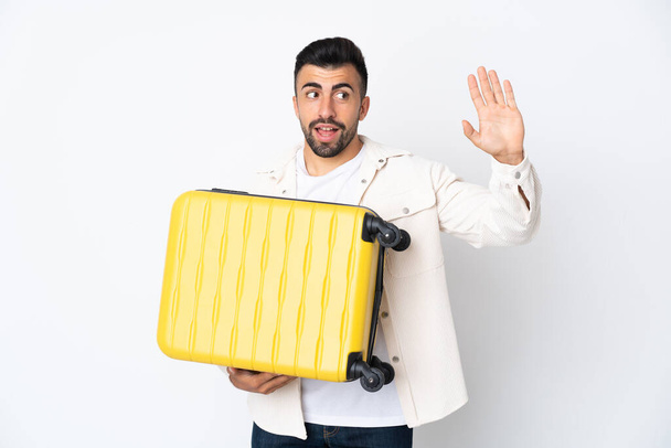 Caucasian man over isolated white background in vacation with travel suitcase and saluting - Φωτογραφία, εικόνα