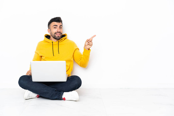 Homem caucasiano sentado no chão com seu laptop apontando o dedo para o lado - Foto, Imagem