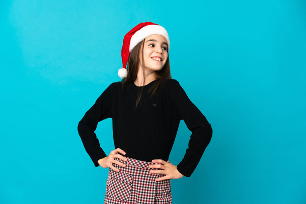 Dziewczynka w świątecznym kapeluszu odizolowana na niebieskim tle, pozująca z rękami na biodrze i uśmiechnięta - Zdjęcie, obraz