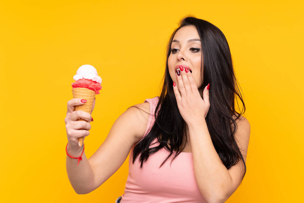 Молода колумбійська дівчина тримає морозиво з корнетом на ізольованому жовтому тлі з подивом і шокованим виразом обличчя. - Фото, зображення