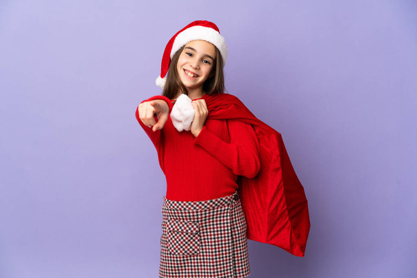 Little girl with hat and Christmas sack isolated on purple background pointing front with happy expression - Foto, afbeelding