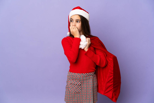 klein meisje met hoed en kerst zak geïsoleerd op paarse achtergrond met twijfels - Foto, afbeelding