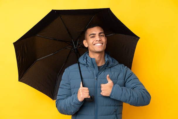 Young handsome man holding an umbrella over isolated yellow background with thumbs up because something good has happened - Photo, Image