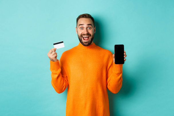Verkkokaupoissa. Innoissaan mies näyttää mobiili näyttö ja luottokortti, hymyillen hämmästynyt, seisoo turkoosi tausta - Valokuva, kuva