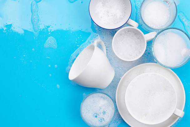 Washing used drinking glasses and cups on wet blue background with soapy foam - 写真・画像