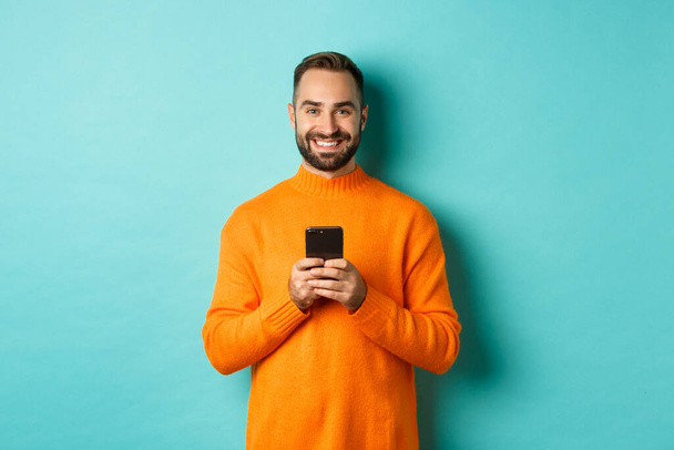 Feliz hombre guapo escribiendo mensaje en el teléfono móvil, sosteniendo el teléfono inteligente y sonriendo, de pie sobre fondo turquesa - Foto, imagen