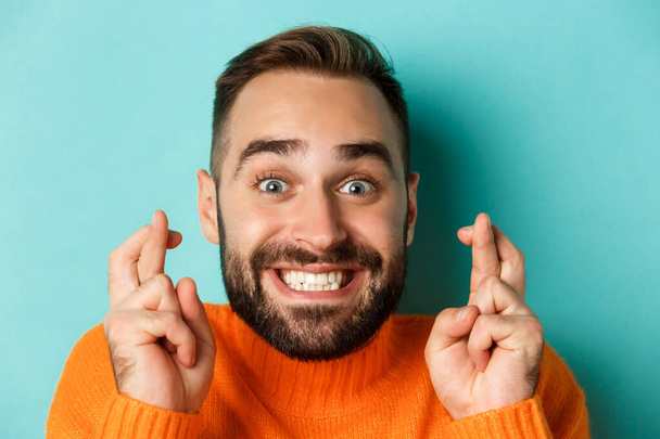 Headshot de homem barbudo esperançoso fazendo um desejo, sorrindo e segurando os dedos cruzados para boa sorte, de pé sobre fundo azul claro - Foto, Imagem