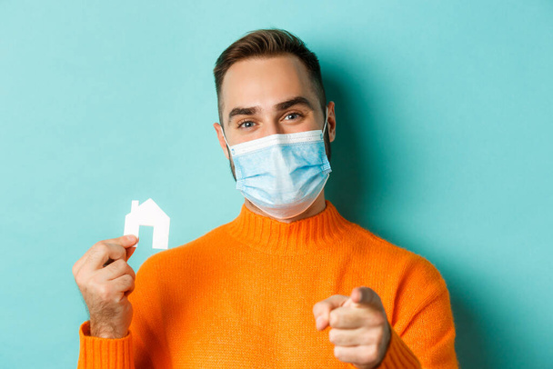 Real estate and coronavirus pandemic concept. Close-up of adult man in medical mask pointing at you and showing small paper house, standing over light blue background - 写真・画像