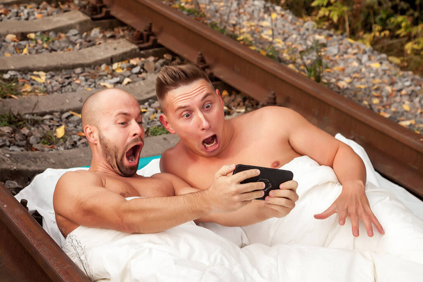 Young emotional gay couple in bed on railway tracks looking at phone. Non-traditional sexual orientation in nature. Concept problem banning gays and not being able to move. Stay at home. Copy space - Foto, Imagen