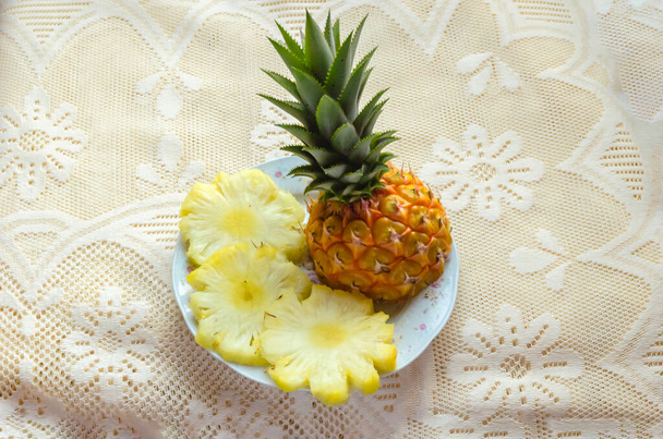 On a lacey tablecloth background, on a floral plate are slices of pineapple and a piece of the fruits in its skin with its green prickly crown on. - Photo, image