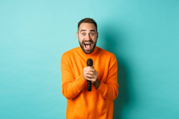Handsome adult man perform song, singing into microphone, standing against turquoise background - Фото, изображение