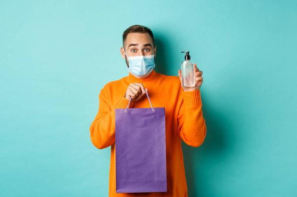 Coronavirus, salgın hastalık ve yaşam tarzı konsepti. Yüz maskeli bir adam alışveriş çantası ve el dezenfektanı gösteriyor, turkuaz arka planda duruyor. - Fotoğraf, Görsel