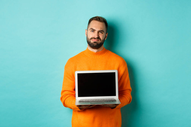 Hezký vousatý muž v oranžovém svetru ukazující obrazovku notebooku, demonstrující promo, šklebící se zklamaně a rozrušeně stojící nad světle modrým pozadím - Fotografie, Obrázek