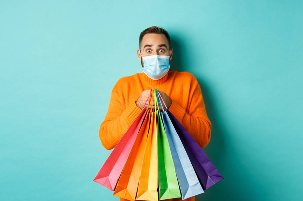 Covid-19, pandemic and lifestyle concept. Excited man in face mask showing shopping bags and rejoicing from discounts, standing over turquoise background - 写真・画像