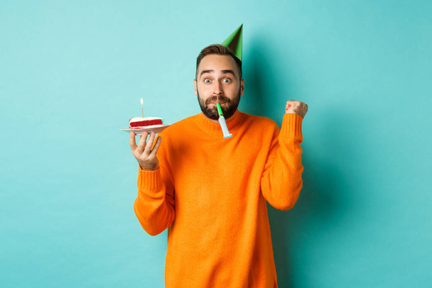 Happy birthday guy celebrating, wearing party hat, blowing wistle and holding bday cake and doing fist pump excited, standing over turquoise background - Photo, Image
