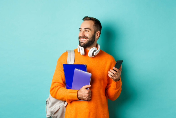 Освіта. Красивий студент чоловічої статі з навушниками та рюкзаком, використовуючи мобільний телефон та тримаючи блокноти, усміхнений щасливий, стоїть над бірюзовим тлом
 - Фото, зображення
