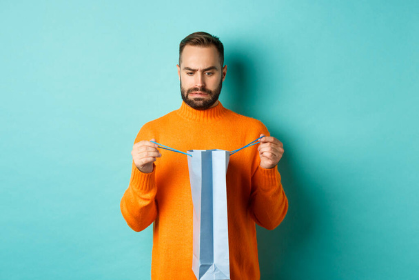 Uomo deluso aperto shopping bag e regalo antipatia, accigliato dispiaciuto, in piedi in maglione arancione contro sfondo turchese - Foto, immagini