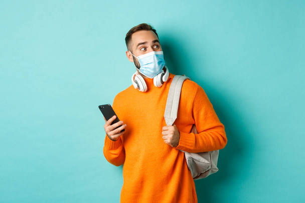 Young man in face mask using mobile phone, holding backpack, staring right amazed, standing against light blue background - Photo, Image