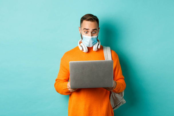 Happy young man in medical mask, working on laptop, looking shocked at computer screen, standing over light blue background - Foto, imagen