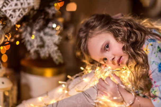 A beautiful girl with curly long hair is sitting near a Christmas tree. Holding a garland of Christmas lights. The concept of winter recreation at home. - Fotografie, Obrázek