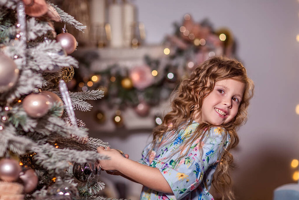 A beautiful laughing girl with curly long hair stands near a decorated Christmas tree. Decorates the Christmas tree. Hangs balloons and toys on the Christmas tree. The concept of winter recreation at home. Close up. - Fotó, kép