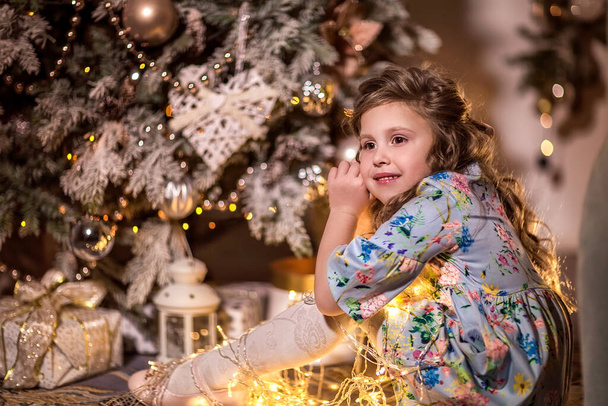A beautiful girl with curly long hair is sitting near a Christmas tree. Holding a garland of Christmas lights. The concept of winter recreation at home. - Fotografie, Obrázek