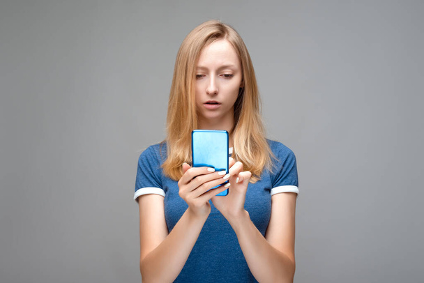 ブロンドの髪と青のTシャツを持つ乱れた女の子は、問題の顔の表情を持っています,スマートフォンを保持しています,何が間違って理解することができません,メッセージの奇妙な意味 - 写真・画像
