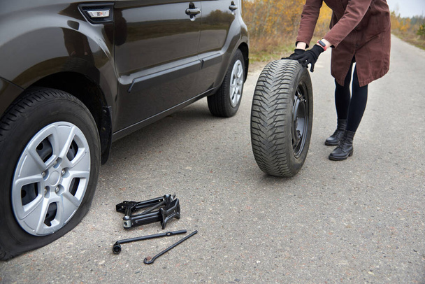 Une jeune femme roule un pneu de secours près de sa voiture avec un pneu crevé, des problèmes sur la route. - Photo, image
