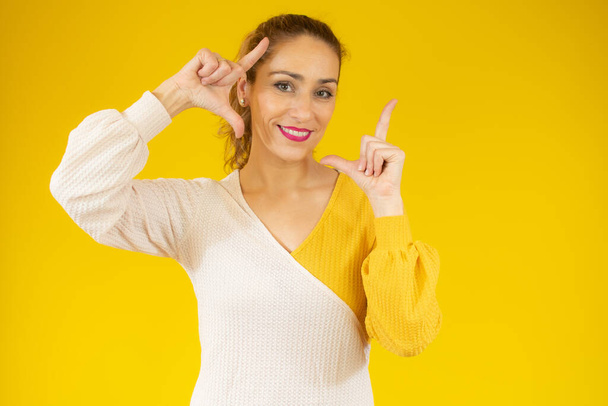 Young beautiful caucasian woman wearing casual sweater standing over isolated yellow background smiling making frame with hands and fingers with happy face. Creativity and photography concept. - Valokuva, kuva