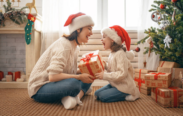 Merry Christmas and Happy Holidays! Cheerful mom and her cute daughter girl exchanging gifts. Parent and little child having fun near tree indoors. Loving family with presents in room.    - Valokuva, kuva