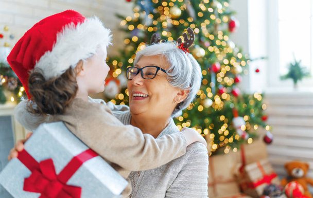 Merry Christmas and Happy Holidays! Cheerful grandma and her cute grand daughter girl exchanging gifts. Granny and little child having fun near tree indoors. Loving family with presents in room. - Zdjęcie, obraz