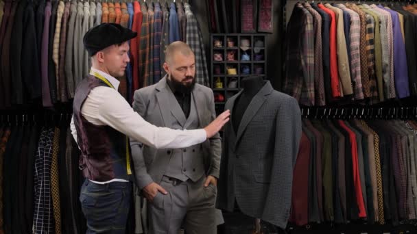アトリエでテーラーメイドでスーツデザインを選ぶハンサムな男のスローモーション。デザイナーが洋服屋のバイヤーにメネキンの衣装を提示し - 映像、動画