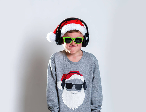 Χαρούμενο αστείο παιδικό πορτραίτο. Δροσερό και έμπιστο παιδί με γυαλιά ηλίου και πουκάμισο Αϊ Βασίλη που κρατάει κουτί δώρου. - Χριστούγεννα. Καλά Χριστούγεννα και Πρωτοχρονιά. Μαθητής με ακουστικά ακούει μουσική.. - Φωτογραφία, εικόνα