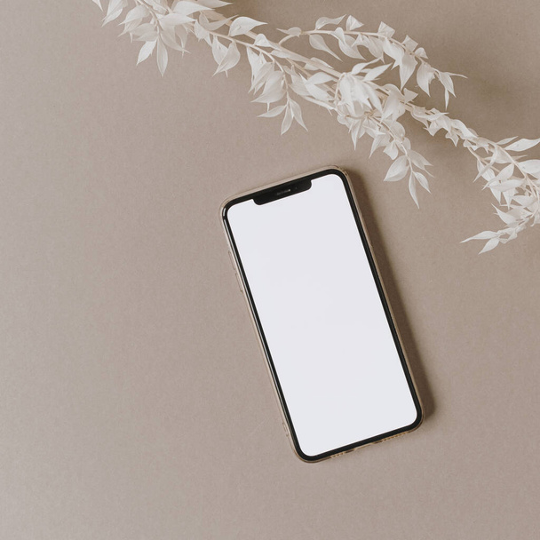 Prázdná obrazovka mobilní telefon, bílá květinová větev na neutrálním pastelově béžovém pozadí. Flat lay, top view minimalistický životní styl blog, webové stránky šablony. Kopírovat mezeru. - Fotografie, Obrázek