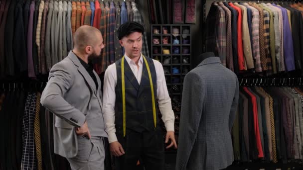 アトリエでテーラーメイドでスーツデザインを選ぶハンサムな男のスローモーション。デザイナーが洋服屋のバイヤーにメネキンの衣装を提示し - 映像、動画
