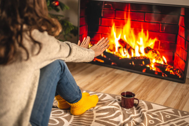 セーターと黄色い暖かい靴下を着た女性が暖炉の前に座っています。これはテレビ画面に描かれ、彼女の手を暖めています。自宅で居心地の良い冬の雰囲気を作成する概念 - 写真・画像