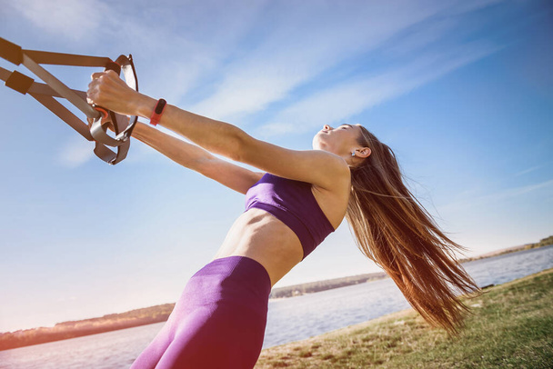 Junge schlanke Frau beim Aufhängungstraining mit Fitnessgurten am Tag im Freien am See. Gesunder Lebensstil - Foto, Bild